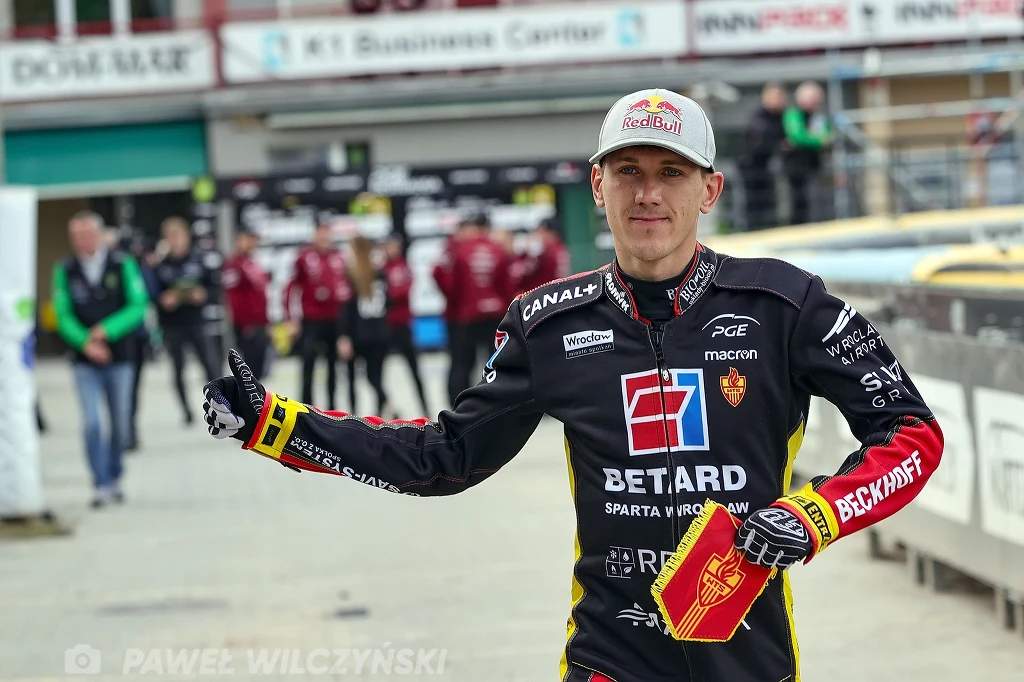 Zawodnik żużlowy w stroju drużynowym z licznymi logotypami sponsorów trzyma kask w ręce i pokazuje gest kciuka do góry na stadionie żużlowym, w tle widocznych jest kilka osób związanych z obsługą imprezy.